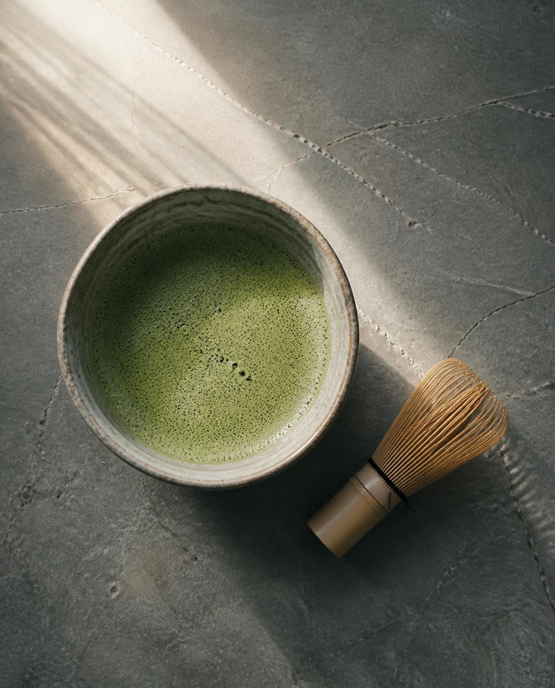 A bowl of whisked matcha beside a bamboo whisk on grey stone, in early light
